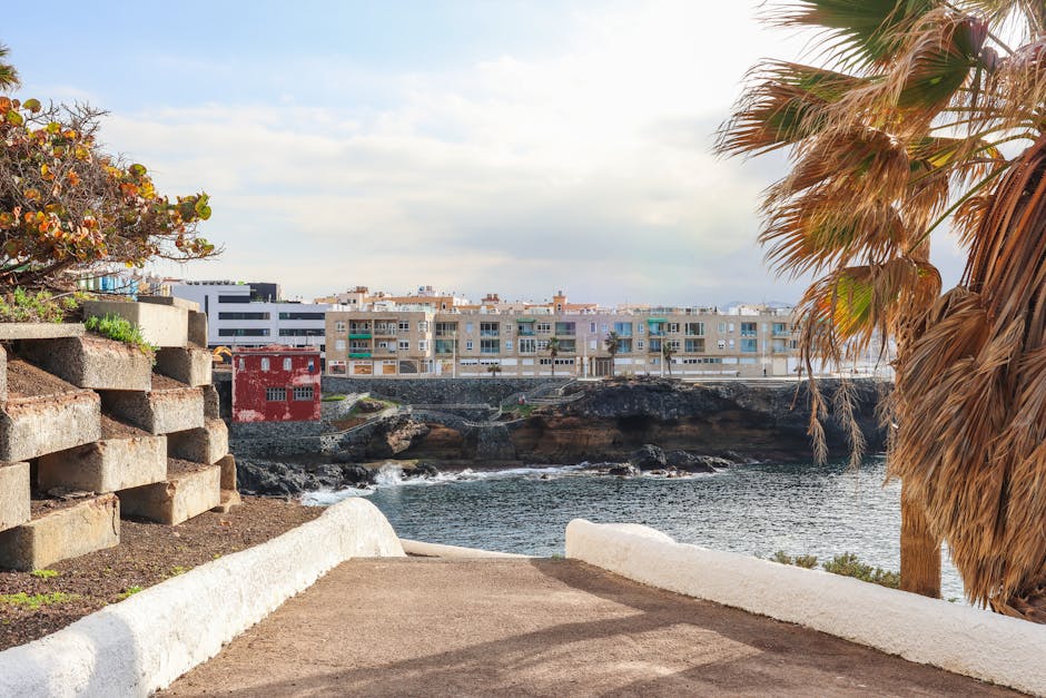 Scenic coastal view from a pathway in Las Palmas de Gran Canaria, Spain, featuring oceanside buildings and palm trees.