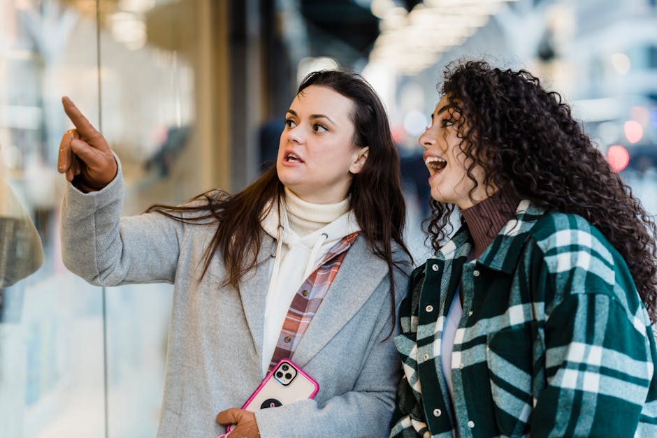 Cheerful multiracial female friends pointing at glass shop window while standing near store on street in city on blurred background