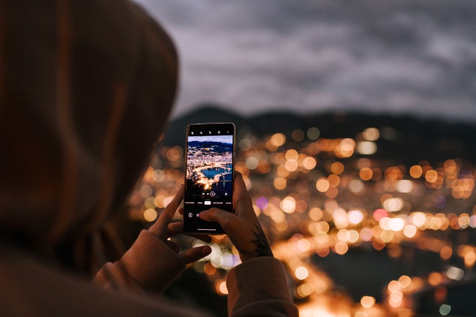 From behind crop person in hoodie taking picture on mobile phone of scenic night city located on hillside and illuminated by bright electric lights
