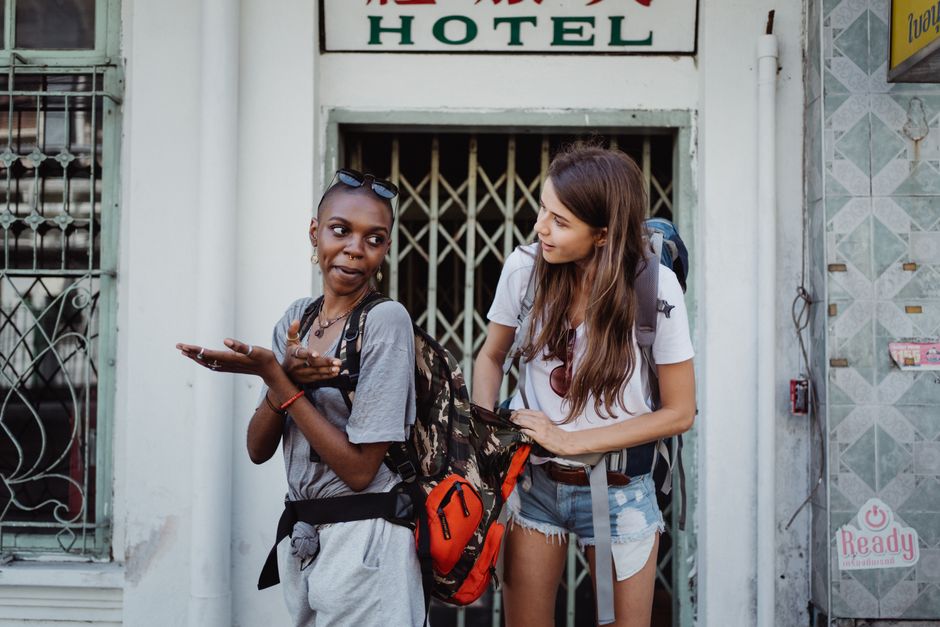 Two women stand laughing with backpacks, exploring a city during their travel adventure.
