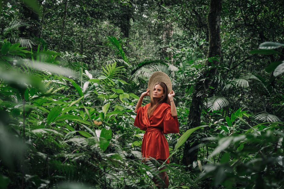 A woman in a vibrant red dress poses among dense forest greenery, creating a captivating nature scene.