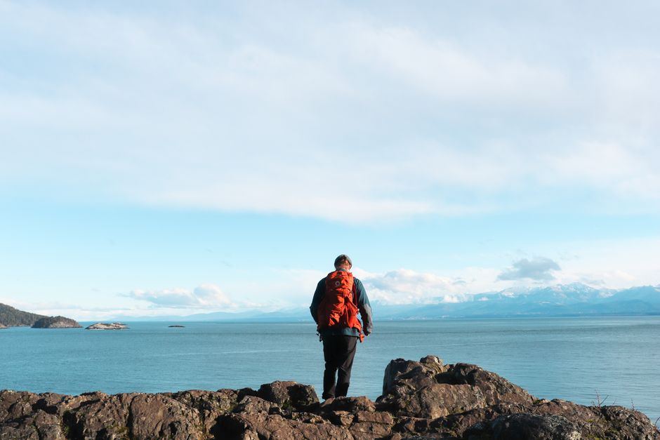 A traveller in an orange backpack stands on rocks gazing at a vast sea and distant mountains.
