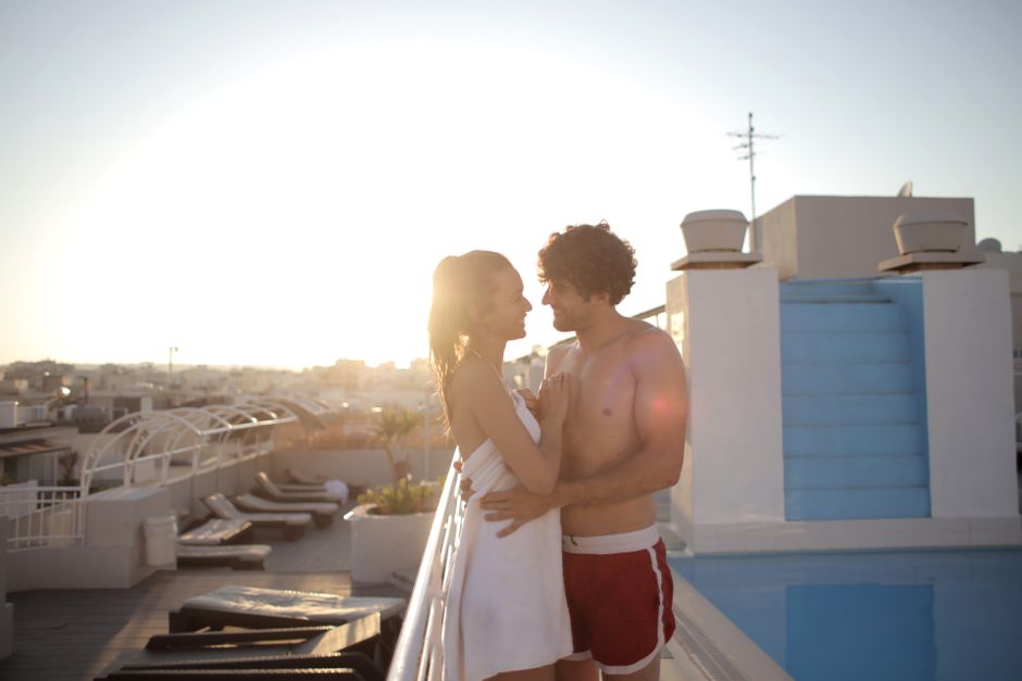 Side view of smiling man in swimwear embracing cheerful beloved in towel while standing near swimming pool at resort in back lit and looking at each other