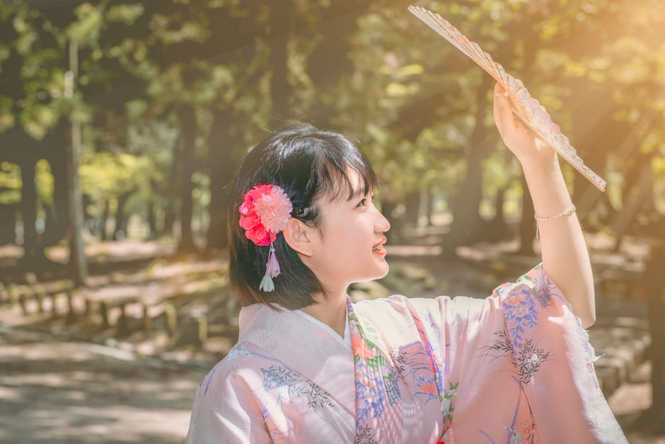 Asian woman in traditional kimono holds a hand fan under sunlight in a serene outdoor setting.