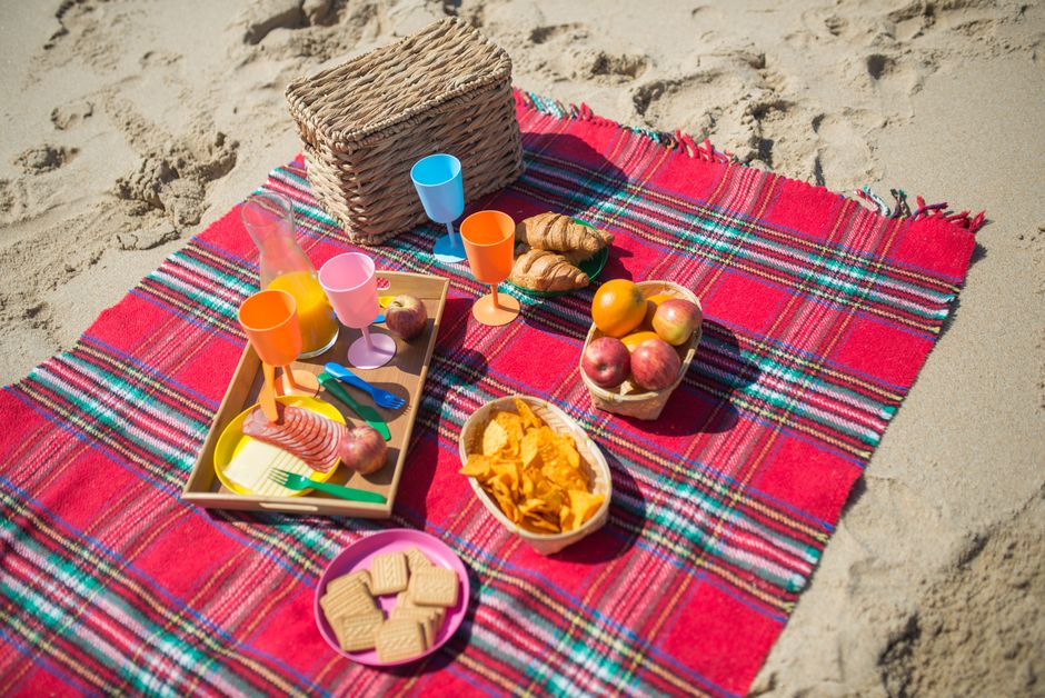 Vibrant beach picnic with fruits, snacks, and drinks on a plaid blanket.
