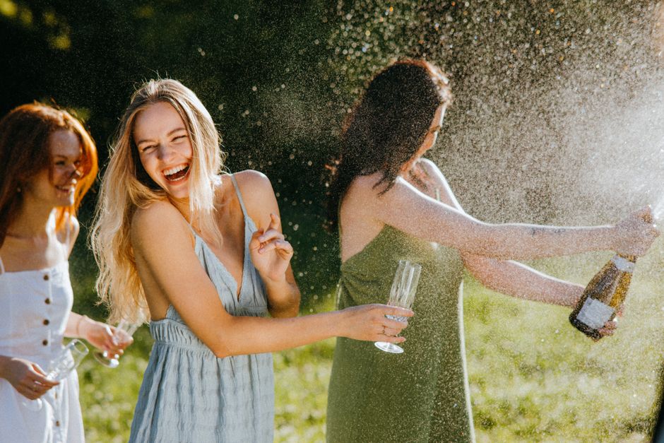 Three women enjoying a sunny day, laughing and celebrating with champagne outdoors.
