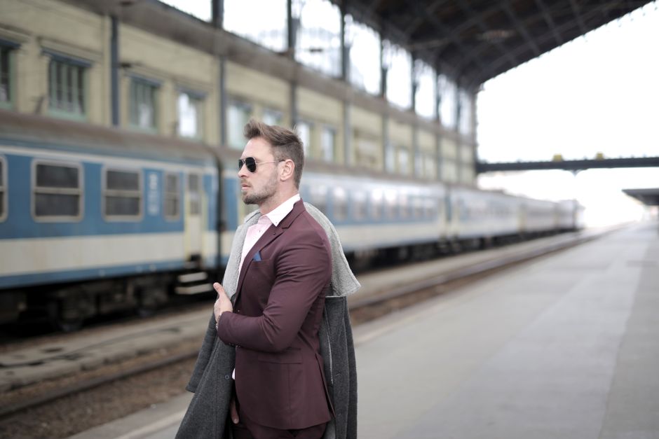 Side view of stylish confident male entrepreneur in formal suit and sunglasses standing on platform with coat on shoulder and waiting for train