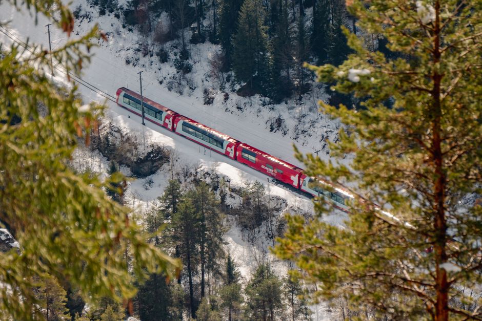 A vibrant red train travels through a snowy winter landscape, surrounded by trees.